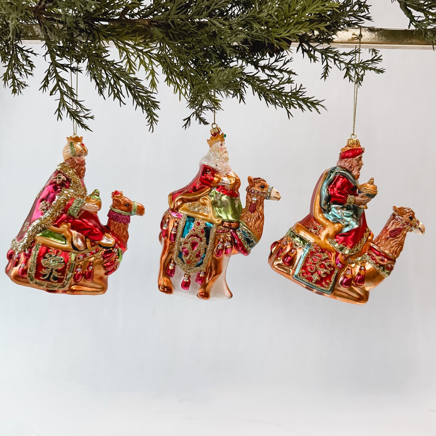 3 threes kings ornaments hanging from a gold bar with greenery against a white background.