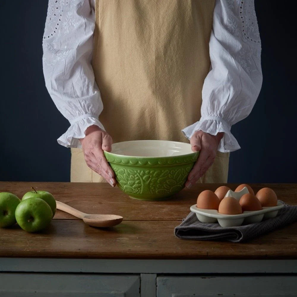 Person holding a green bowl on a wooden table with apples, a wooden spoon, and eggs.