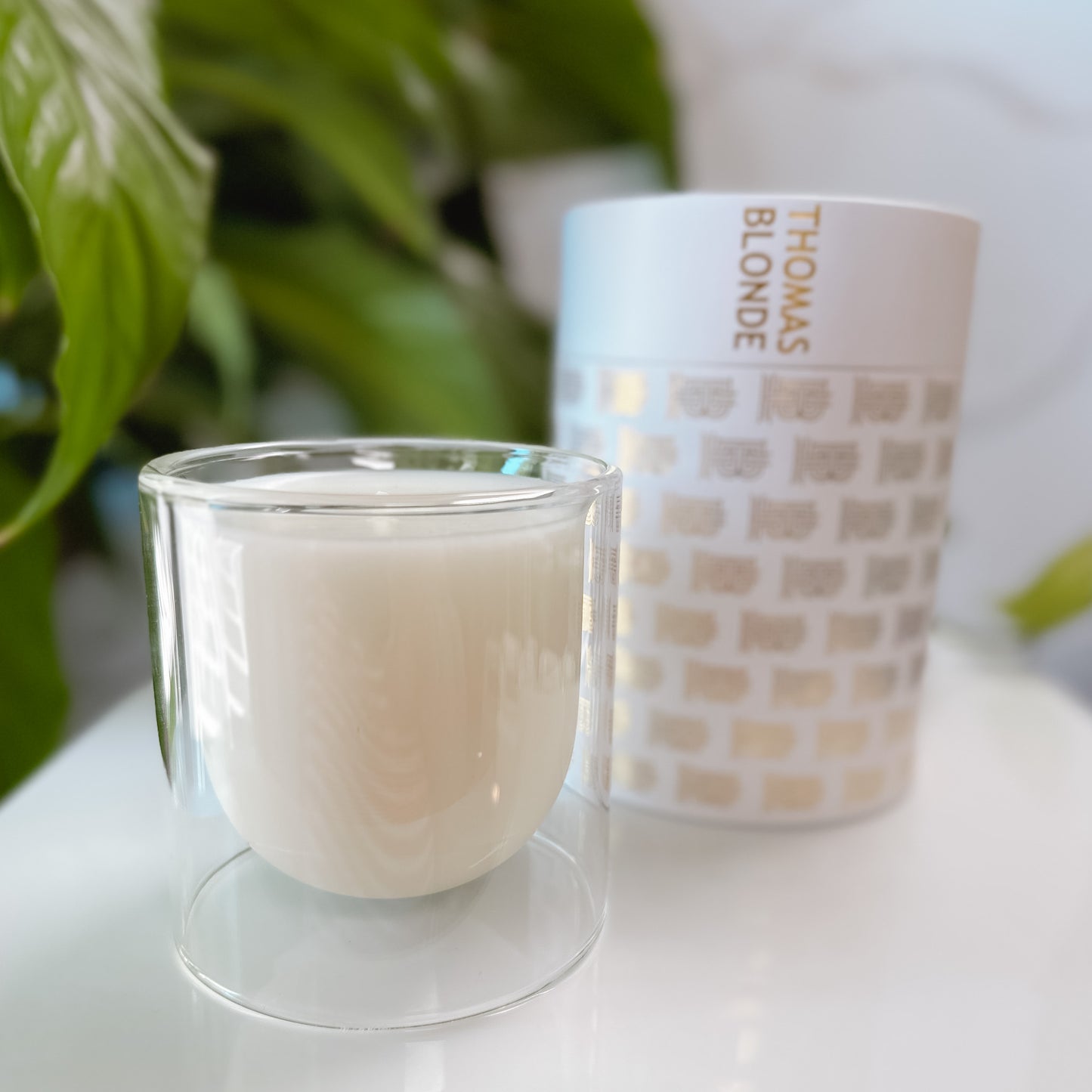Thomas Blonde candle in a clear glass jar with it's packaging on a white tray with foliage in the background.