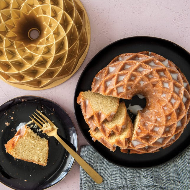 jubilee bundt pan displayed next to a sliced bundt cake on a plate and a napkin on a pale purple backgound