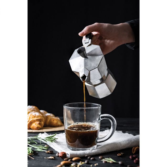 a persons hand pouring coffee out of the aluminum espresso maker into a clear glass mug against a black background