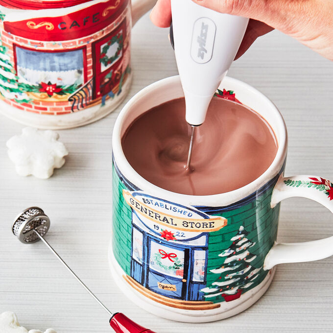 frother at work in a holiday mug mixing beverage with another mug and a cookie in the background