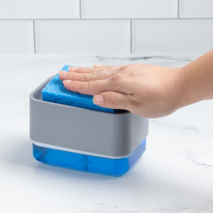 a person using the soap dispensing sponge holder with a sponge while it sits on a white marble countertop