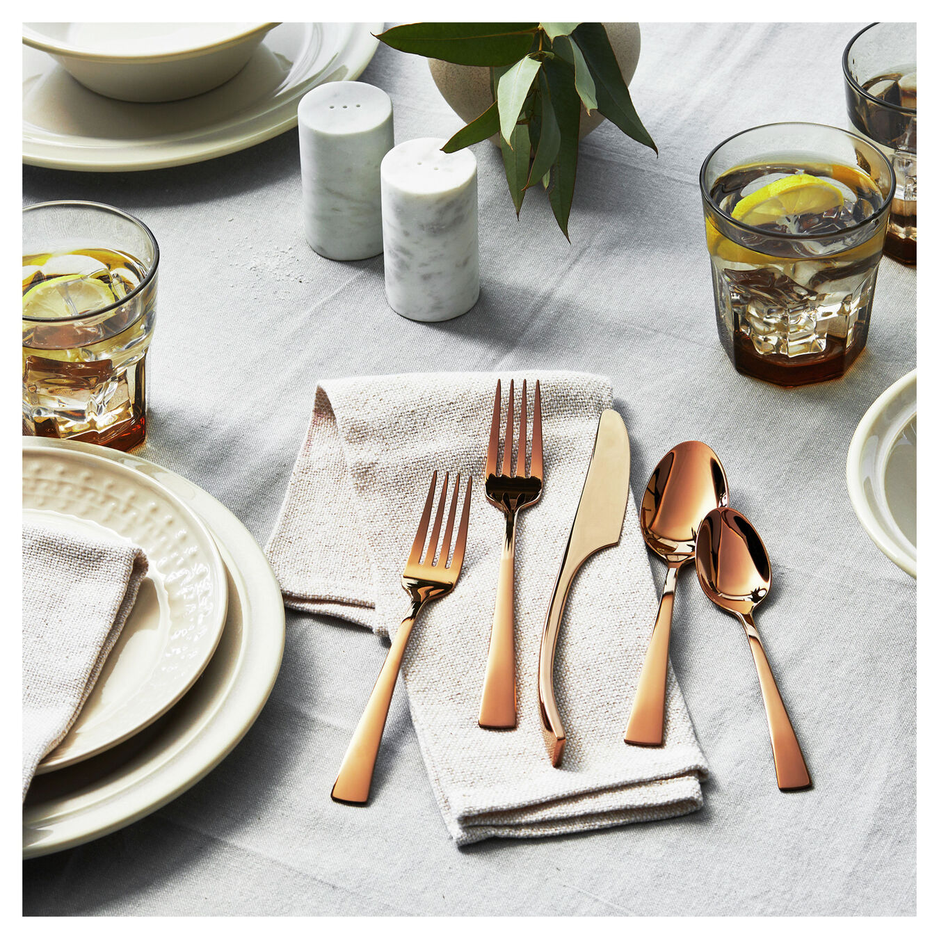 5 pieces of the bellasera gold flatware displayed on a table setting next to dinner plates, salt and pepper shaker, drinking glasses, and a vase with green sprigs on a white tablecloth