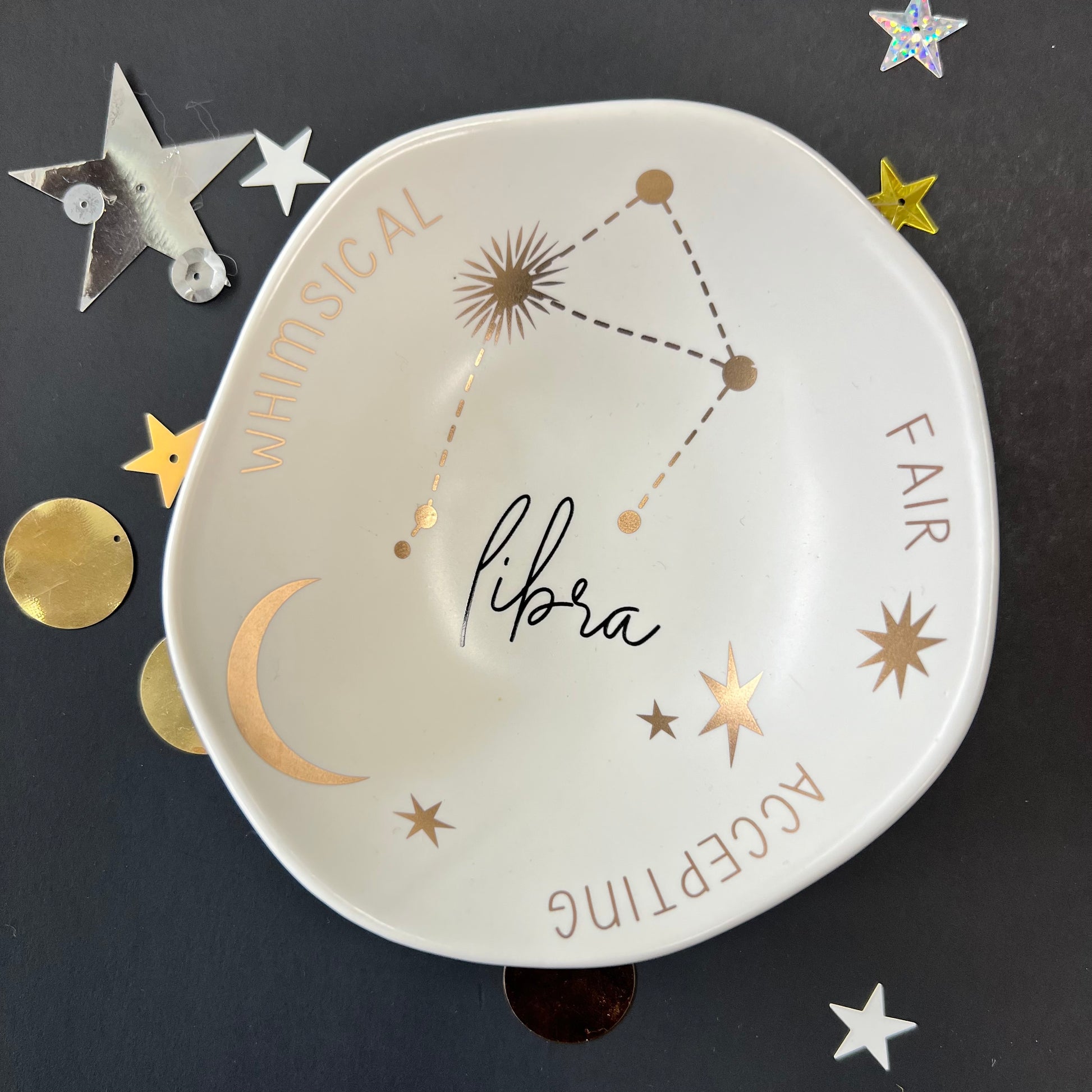 cream dish with gold stars and "Libra Whimsical Fair Accepting" around the inner rim on a black background with scattered stars and orbs.