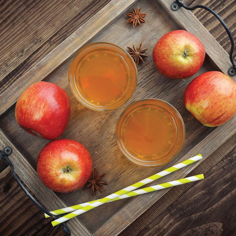 top view of two apple cider drinks displayed on a wooden tray with four apples, star anis, and two straws