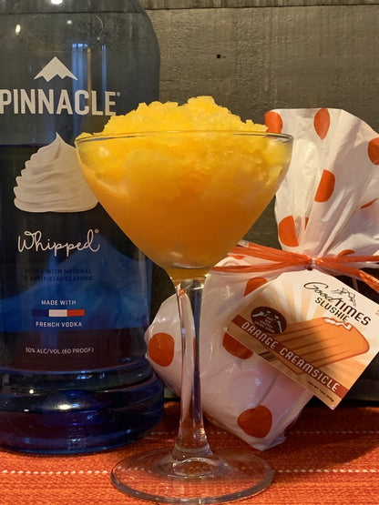 orange creamsicle slushie displayed in a glass next to the package and bottle of vodka on a rust colored tablecloth