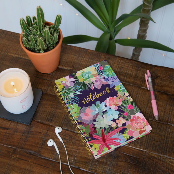 notebook on wooden table with earphones, pen, candle, and plants.