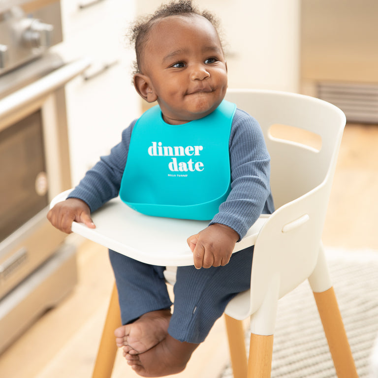 a baby boy sitting in a highchair in the kitchen wearing the dinner date wonder bib