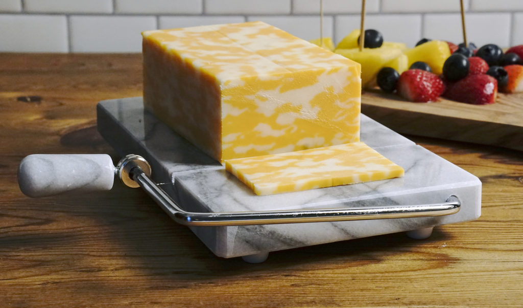 slicer on wood counter with cheese sliced, fruit in background.