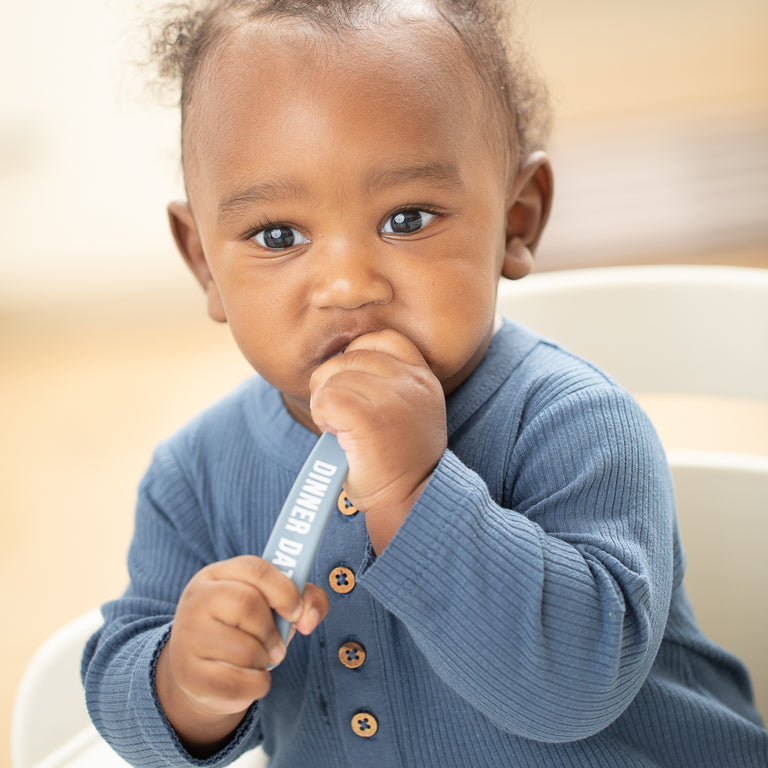 a baby boy sitting in a highchair chewing on the blue spoon