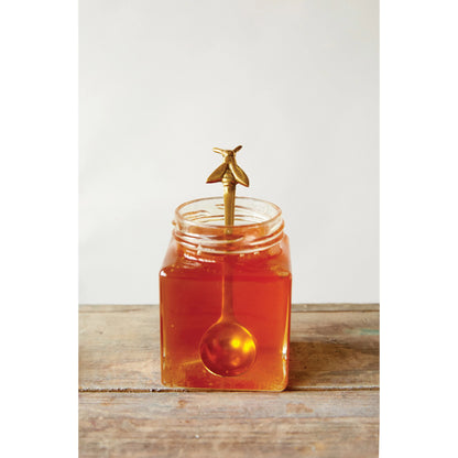 brass spoon with bee displayed in a jar of honey on a rustic table against a white background