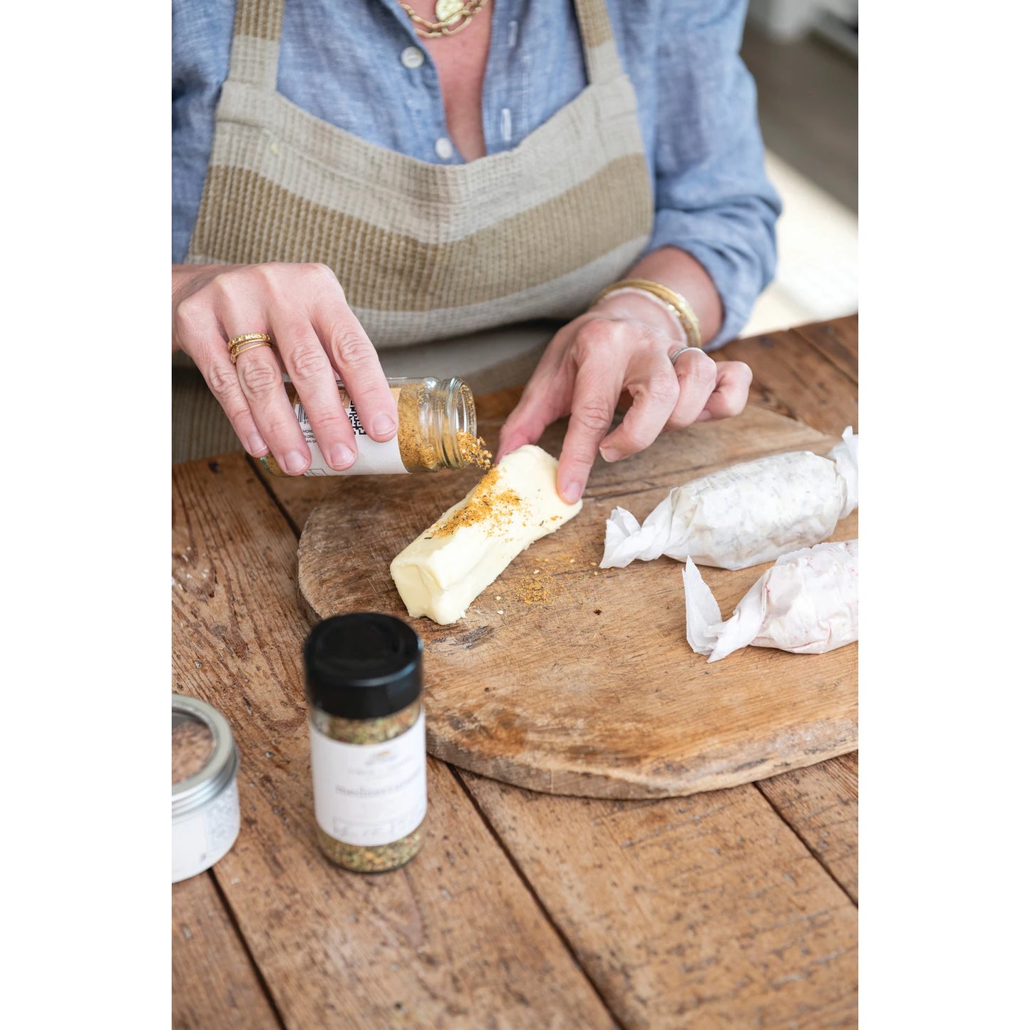 a woman seasoning food with the mediterranean spice blend on a round wooden board sitting next to the jar on a wooden slat table