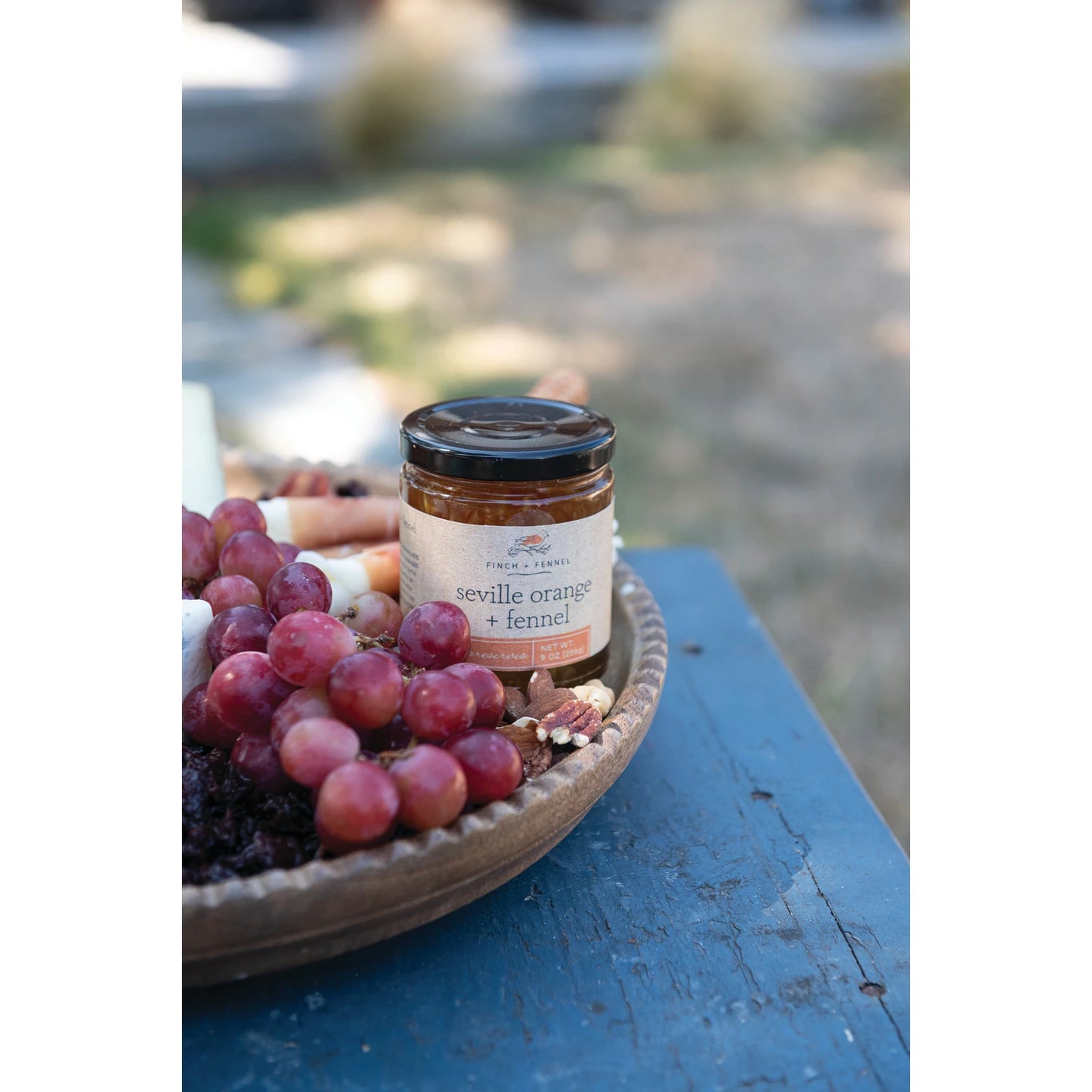 jar of seville orange and fennel displayed on a bowl next to grapes, cheeses, and nuts on a blue table outside