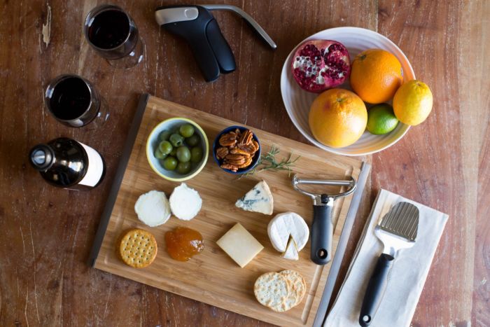 cheese slicer on wood board with various cheeses, jam, crackers, and olives.