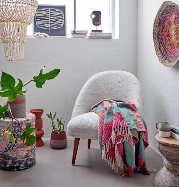 recycled cotton blend striped throw displayed bunched over a chair in the corner of a room next to potted plants, wall hangings, and side table