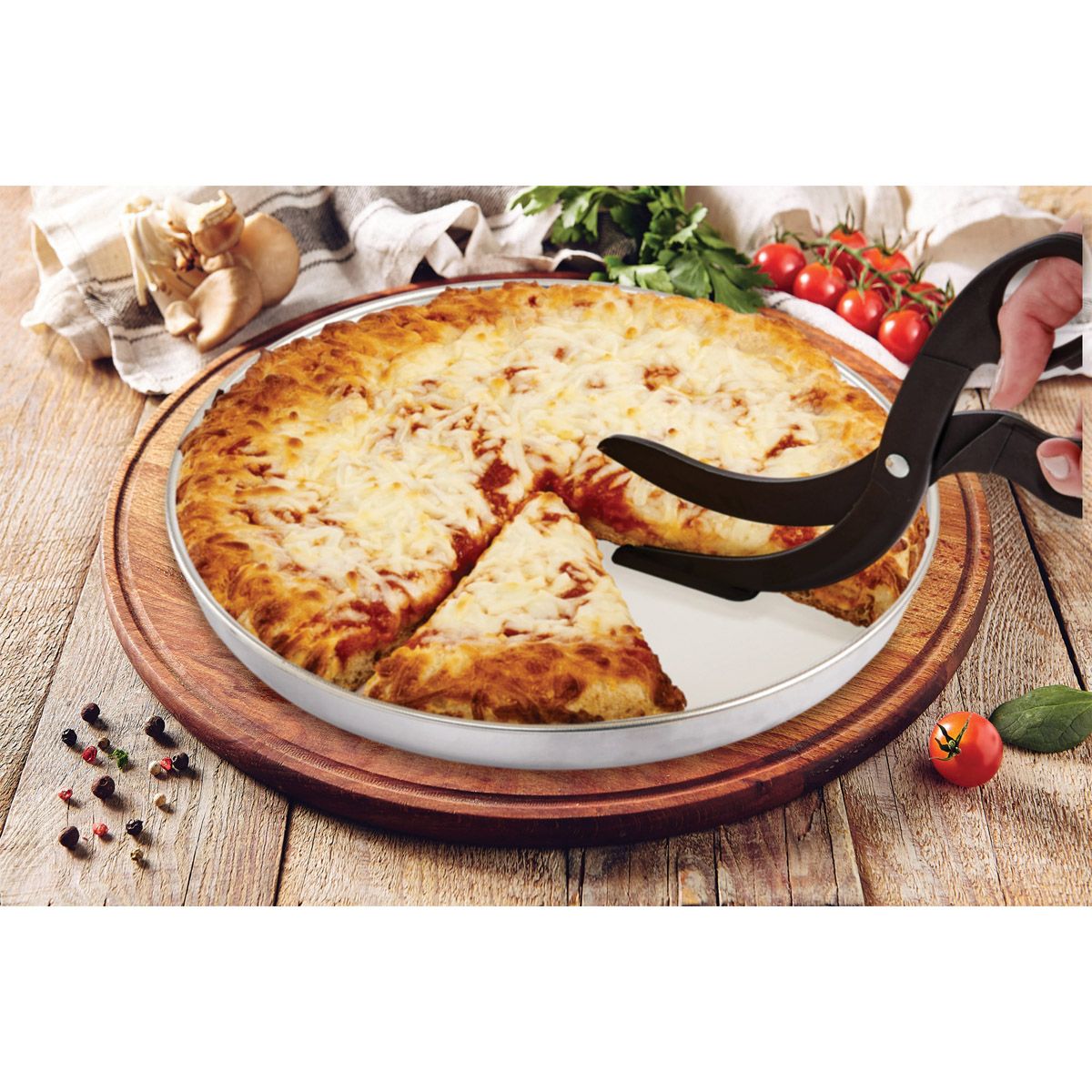 a persons hand illustrating cutting a pizza with the pizza scissors surround by tomatoes on the vine, spices, garlic cloves, and a towel