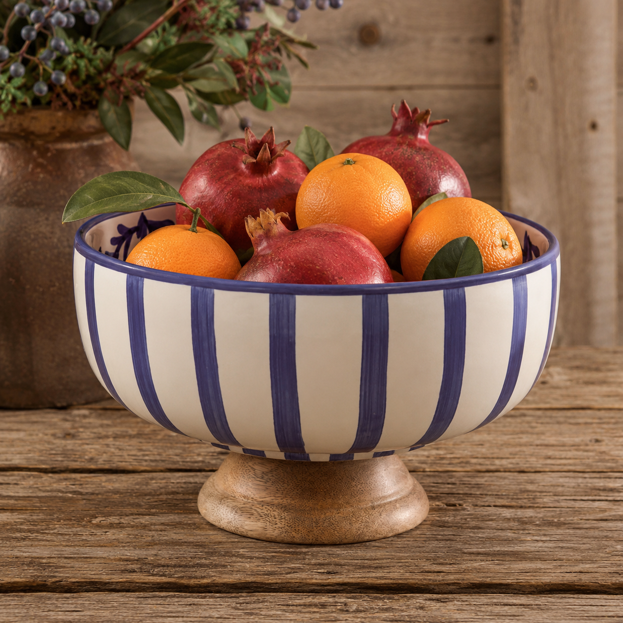 A ceramic bowl with blue and white vertical stripes and a wooden pedestal base, filled with oranges and pomegranates on a rustic wooden table.