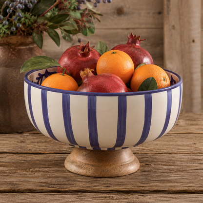 A ceramic bowl with blue and white vertical stripes and a wooden pedestal base, filled with oranges and pomegranates on a rustic wooden table.
