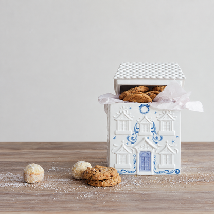 A white ceramic gingerbread house cookie jar with blue detailing, shown open with cookies inside on a wooden surface against a light neutral background.