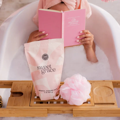 Person in a bathtub with a pink book and 'sweet grace' mineral soaping salts in the foreground