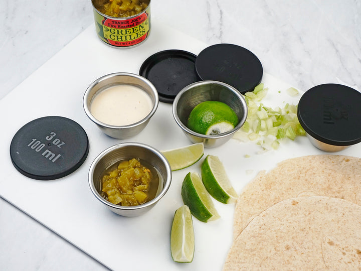 A set of black condiment cups with lids on a kitchen counter, with one cup filled with guacamole, another with salsa, a third with lime slices, and a fourth with lettuce. 