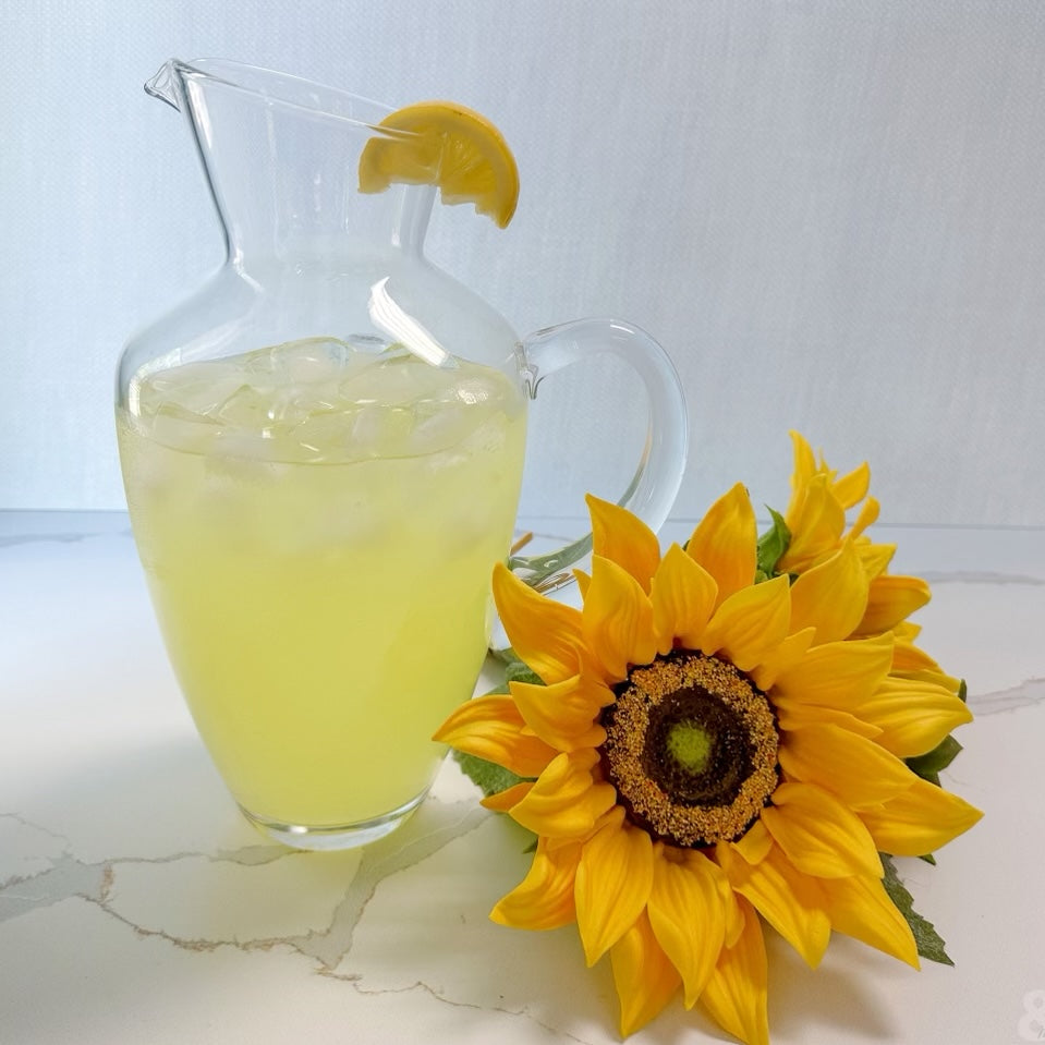 glass pitcher filled with lemonade and a lemon wedge on the rim set on a marble countertop with sunflowers.