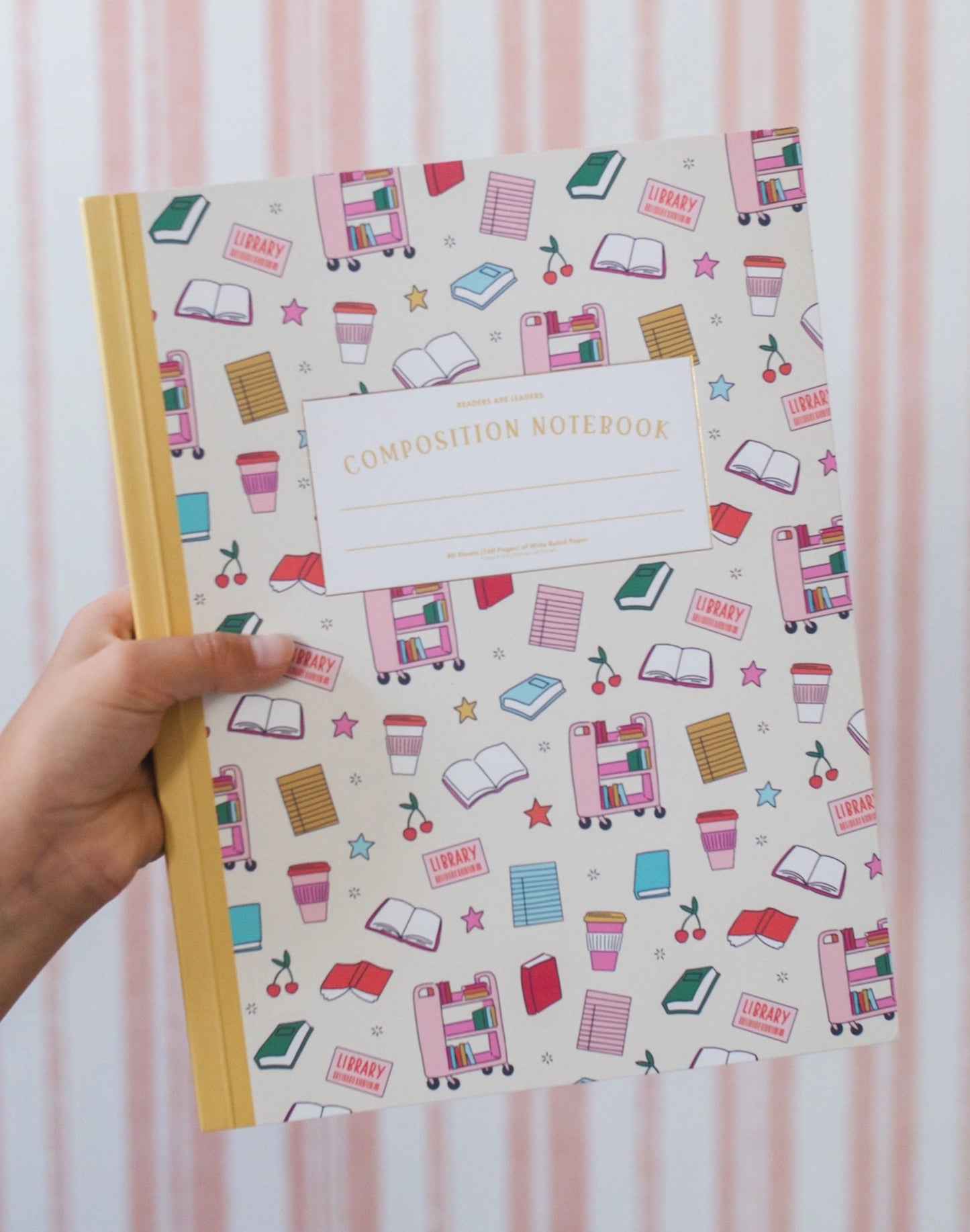 Hand holding a composition notebook with a colorful pattern on a pink striped background