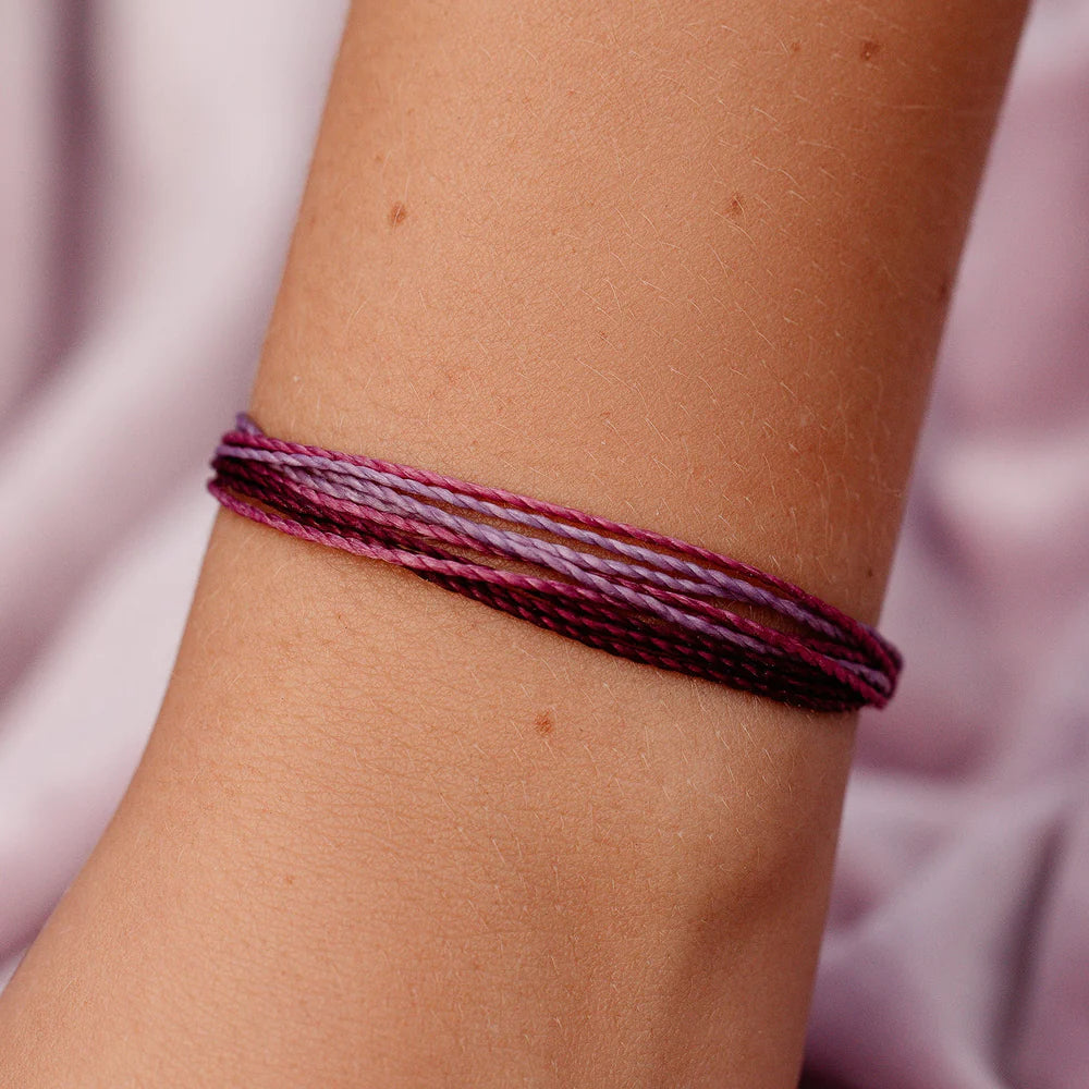 Pura Vida multi strand corded bracelet in varying shades of purple and pink with a Pura Vida logo charm on a wrist