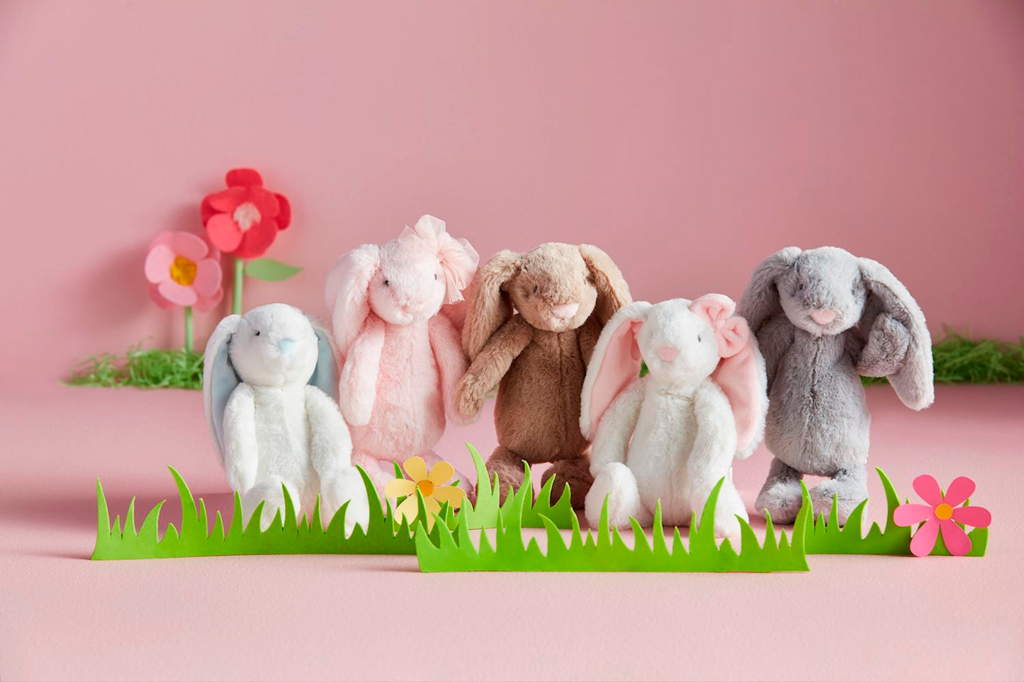 Five plush bunny toys in various colors on a pink background with cut-out paper flowers and grass.
