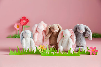 Five plush bunny toys in various colors on a pink background with cut-out paper flowers and grass.
