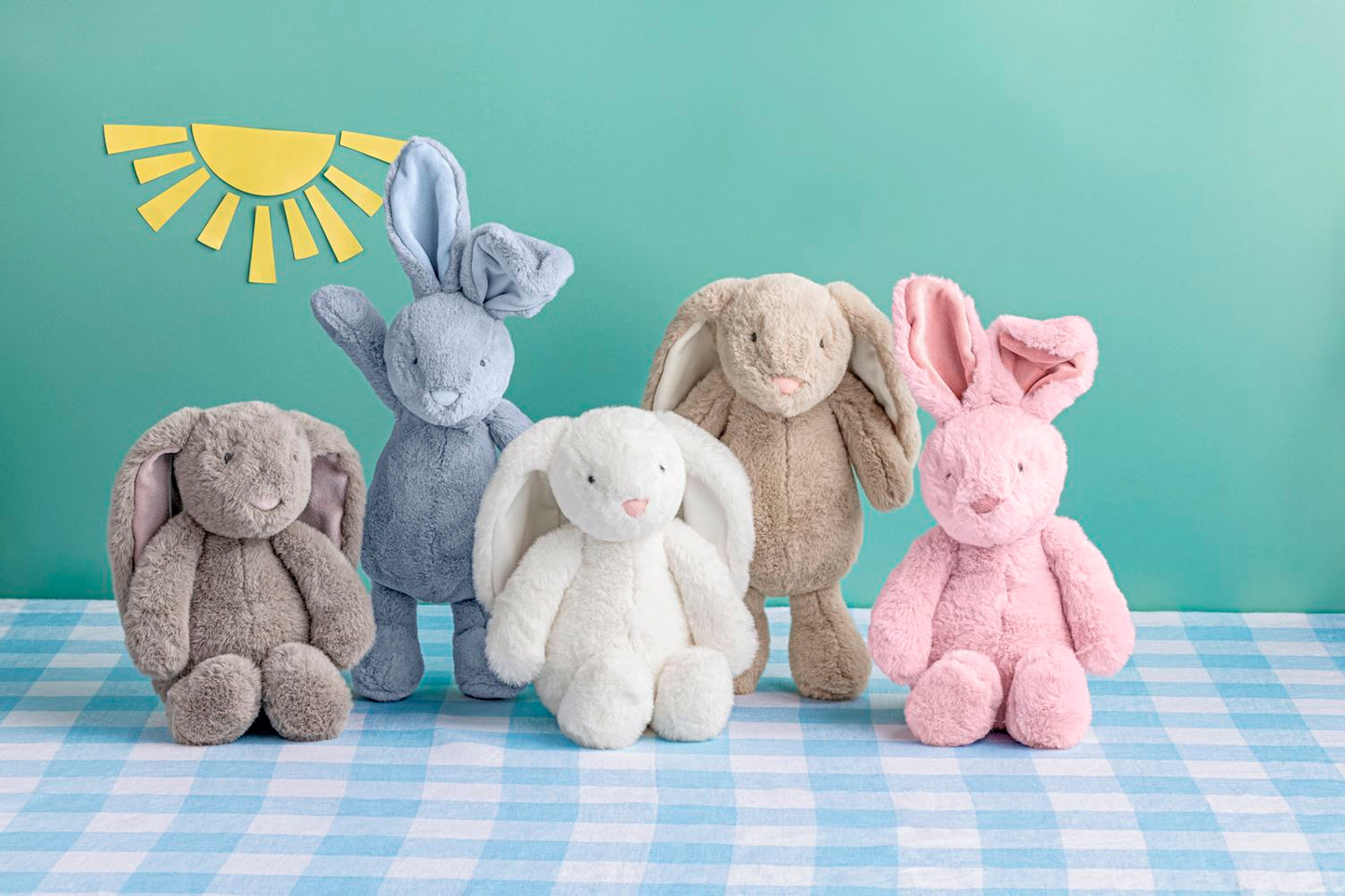 Five plush bunny toys in different colors on a blue checkered tablecloth with a light green background with paper sunshine.