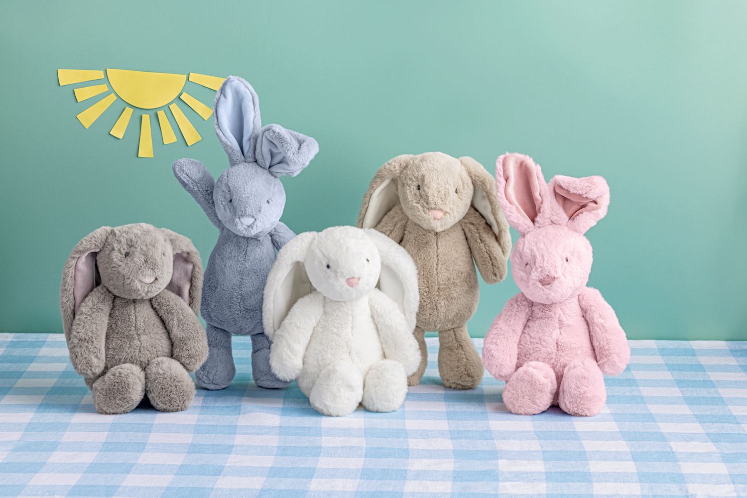 Five plush bunny toys in different colors on a blue checkered tablecloth with a light green background with paper sunshine.