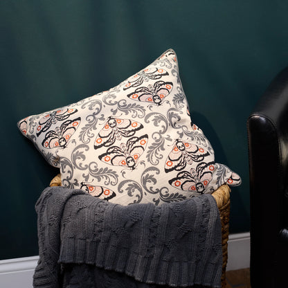 A square throw pillow with a cream background and a detailed moth pattern in black, gray, and orange, styled on a wicker chair with a dark gray knit blanket against a dark green wall.