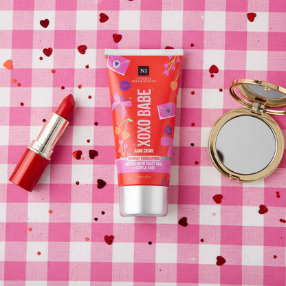 Makeup products including a red lipstick, red tube of XOXO BABE cream, and compact mirror on a pink checkered background with heart shaped confetti.