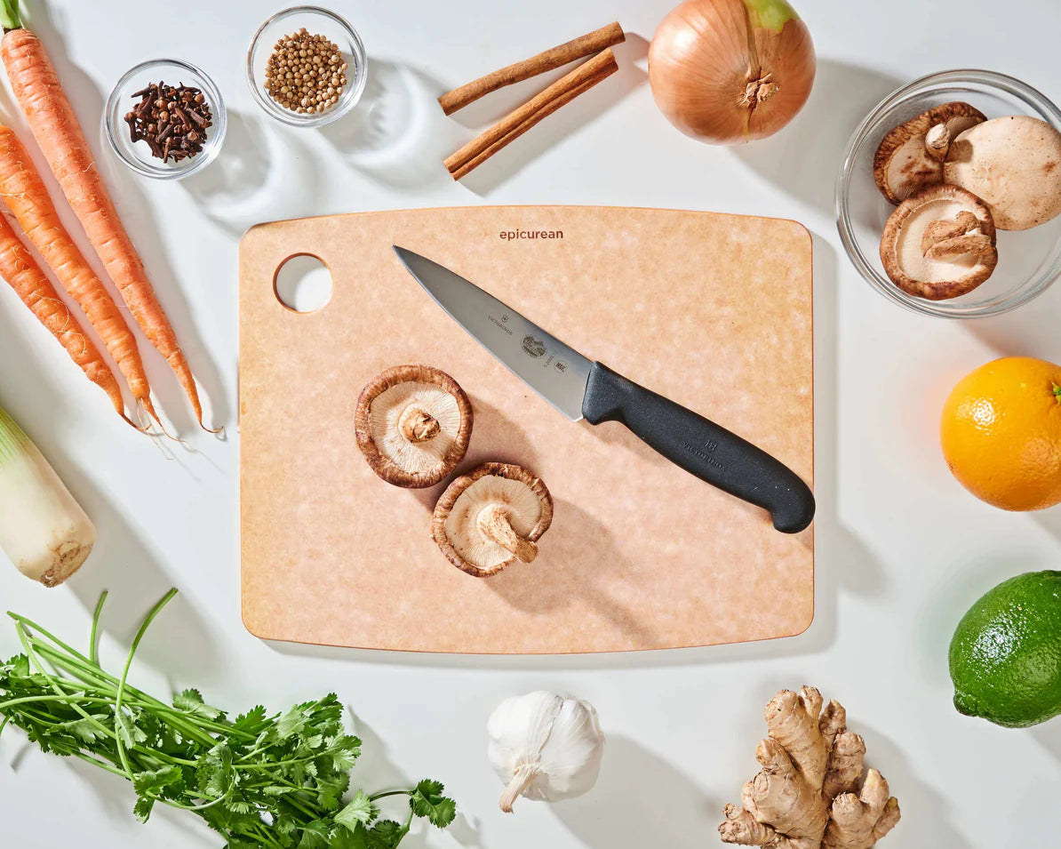 medium natural epicurean cutting board with a knife on it on a countertop with fruit, veggies, and spices.