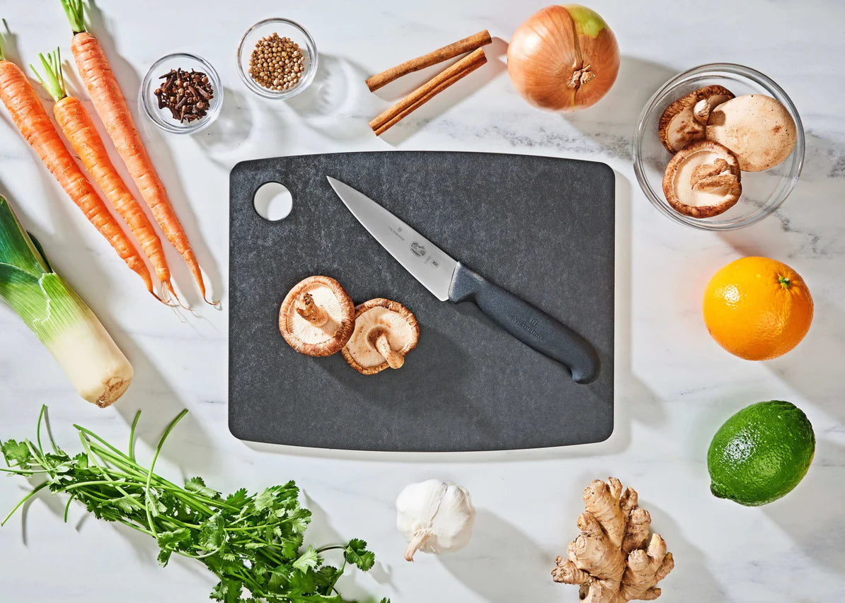 medium slate epicurean cutting board with a knife on it on a countertop with fruit, veggies, and spices.