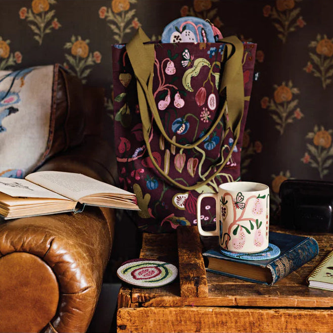 A patterned tote bag stands on a rustic wooden table in a cozy room setting, surrounded by books, a ceramic mug,  and wallpapered walls.
