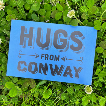 blue postcard with "hugs from conway" printed in black laying in a field of clovers.