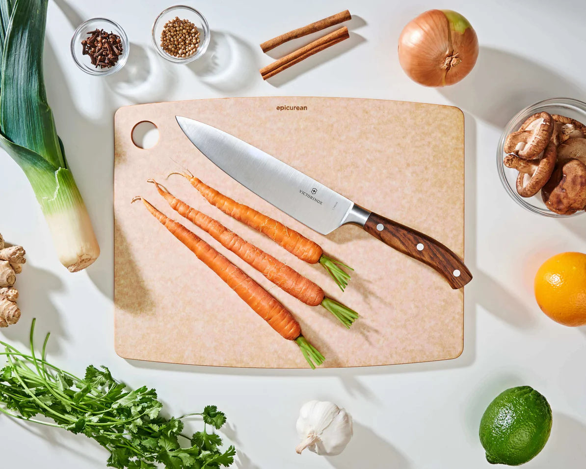 large natural epicurean cutting board with a knife on it on a countertop with fruit, veggies, and spices.