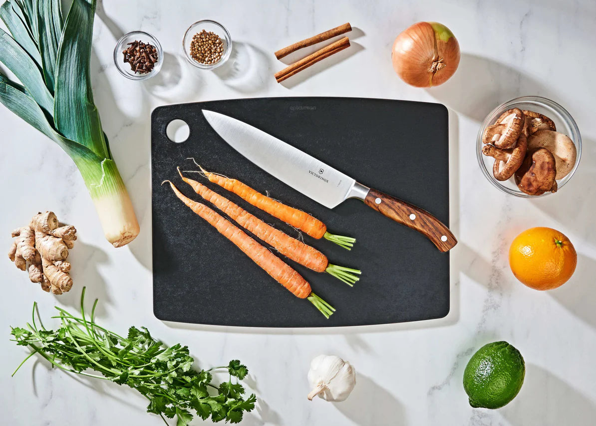 large slate epicurean cutting board with a knife on it on a countertop with fruit, veggies, and spices.