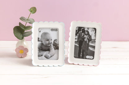 Two white photo frames with family photos set on a white table with a vase and greenery on a pink background