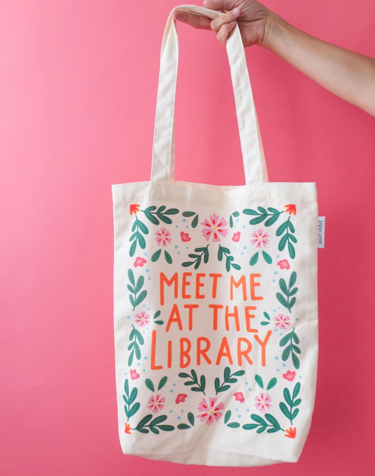 Tote bag with 'Meet Me at the Library' text and floral design on a pink background