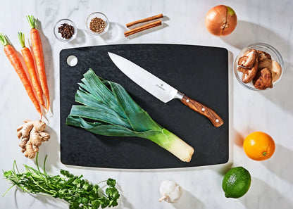 larger slate epicurean cutting board with a knife on it on a countertop with fruit, veggies, and spices.