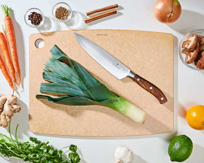 larger natural epicurean cutting board with a knife on it on a countertop with fruit, veggies, and spices.