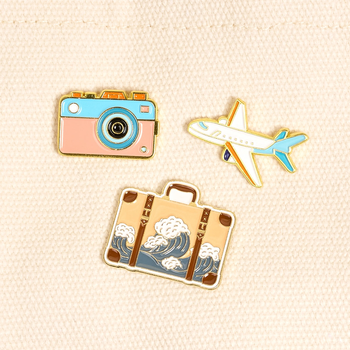 Three travel-themed pins including a camera, airplane, and suitcase on a beige fabric background.
