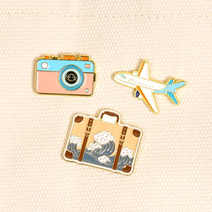 Three travel-themed pins including a camera, airplane, and suitcase on a beige fabric background.
