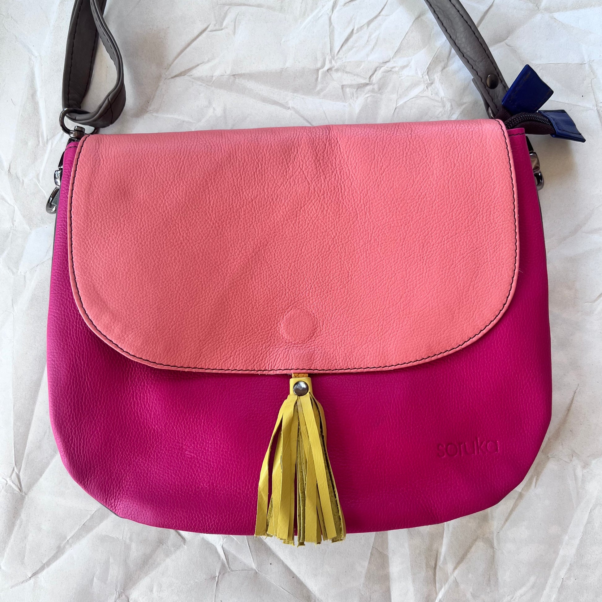 rounded fuchsia purse with pink flap, yellow tassel, and grey strap.