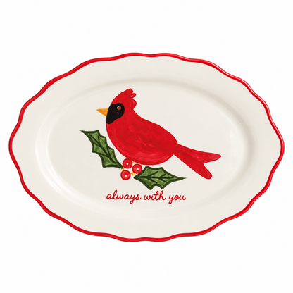 An oval white ceramic serving platter with a scalloped red rim, featuring a painted red cardinal perched on a green holly branch with red berries and the words 'always with you' in red script.