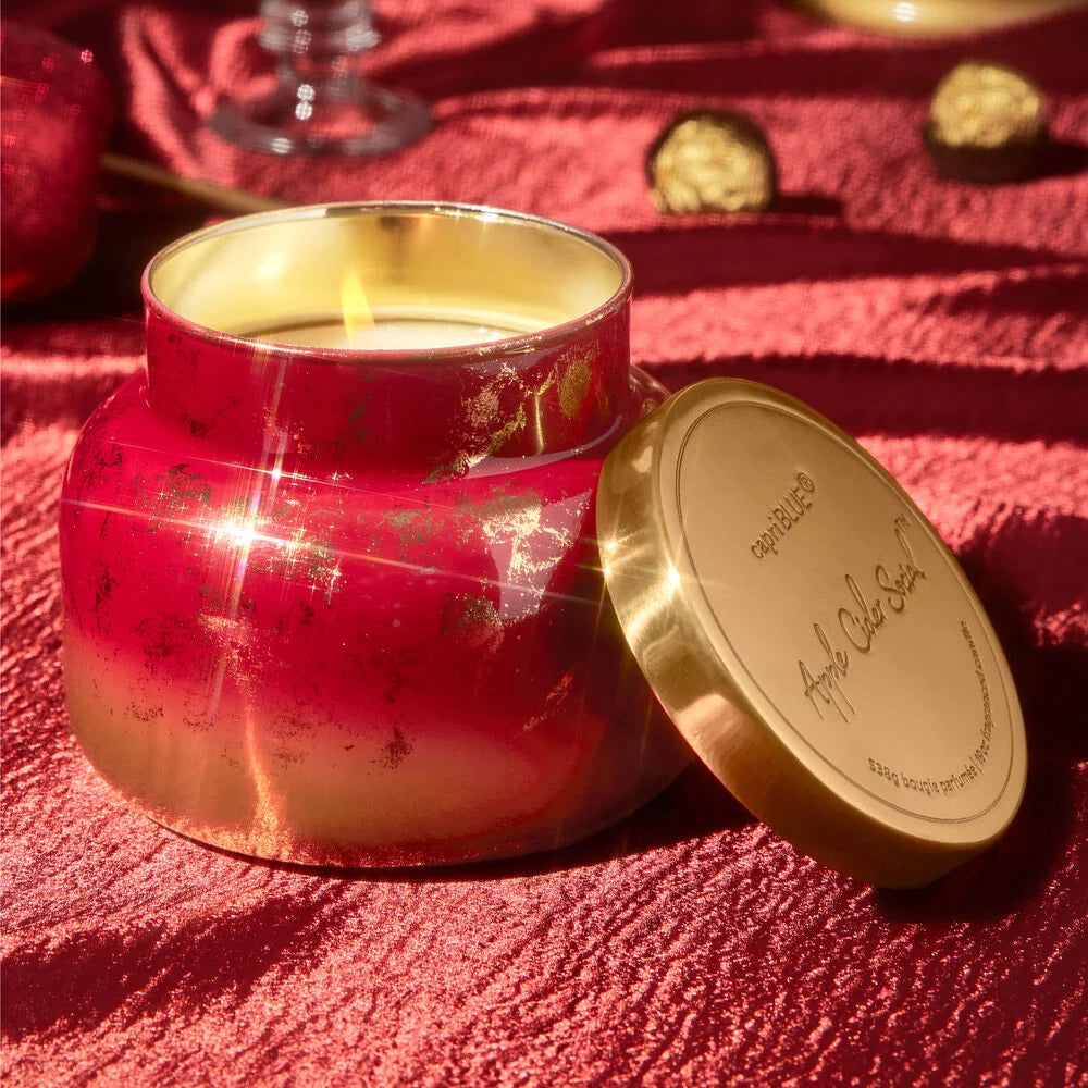 lit Apple Cider Social Glimmer Candle set on a table covered with a red cloth with chocolates and a glass in the background.